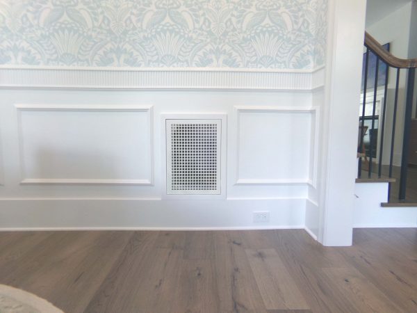 Frontal view of a Wood Return Egg Crate Fitter Grille built into the white painted wainscoting of a dining room with wood floors and light blue wallpaper on the wall above.