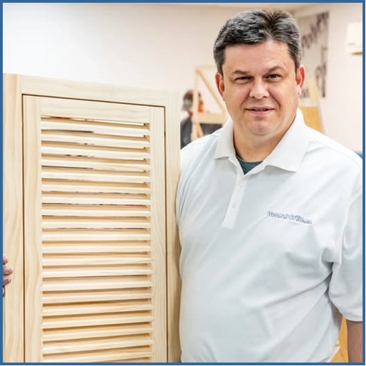 A man, Rod Gunter, is standing in a workshop wearing a white WoodAirGrille logo shirt and holding a wood return air filter grille in his right arm