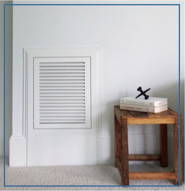 Wood return air filter grille installed in white wall beside a small square wood table. A large jack and two books sit on the table