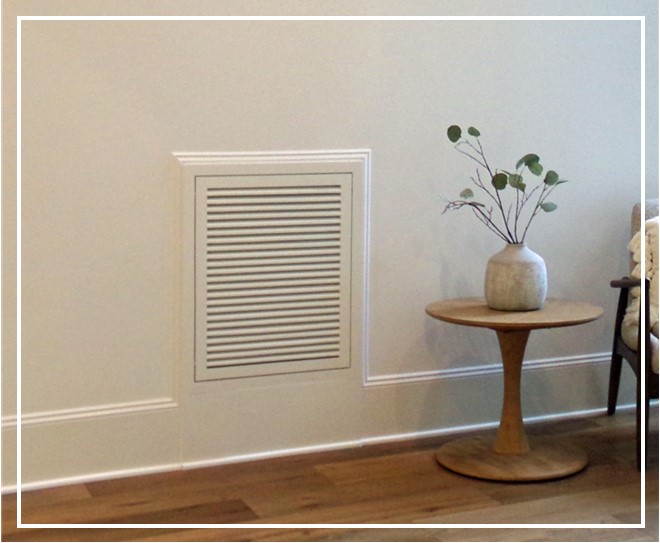 Wood Return Air Filter Grille installed in white wall above baseboard beside small round wood table on wood floors with plant in vase on table