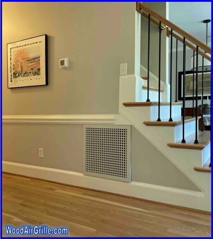 The file "Wood Return Air Grille hero shot.jpg" displays a brightly lit interior hallway featuring warm hardwood floors and light grey walls with crisp white chair rail molding. Prominently featured beneath a staircase is a white, square wood return air grille with a grid pattern, seamlessly integrated into the wall between the baseboard and chair rail. The adjacent staircase has wooden treads, white risers, and black metal balusters. A framed artwork hangs on the left.